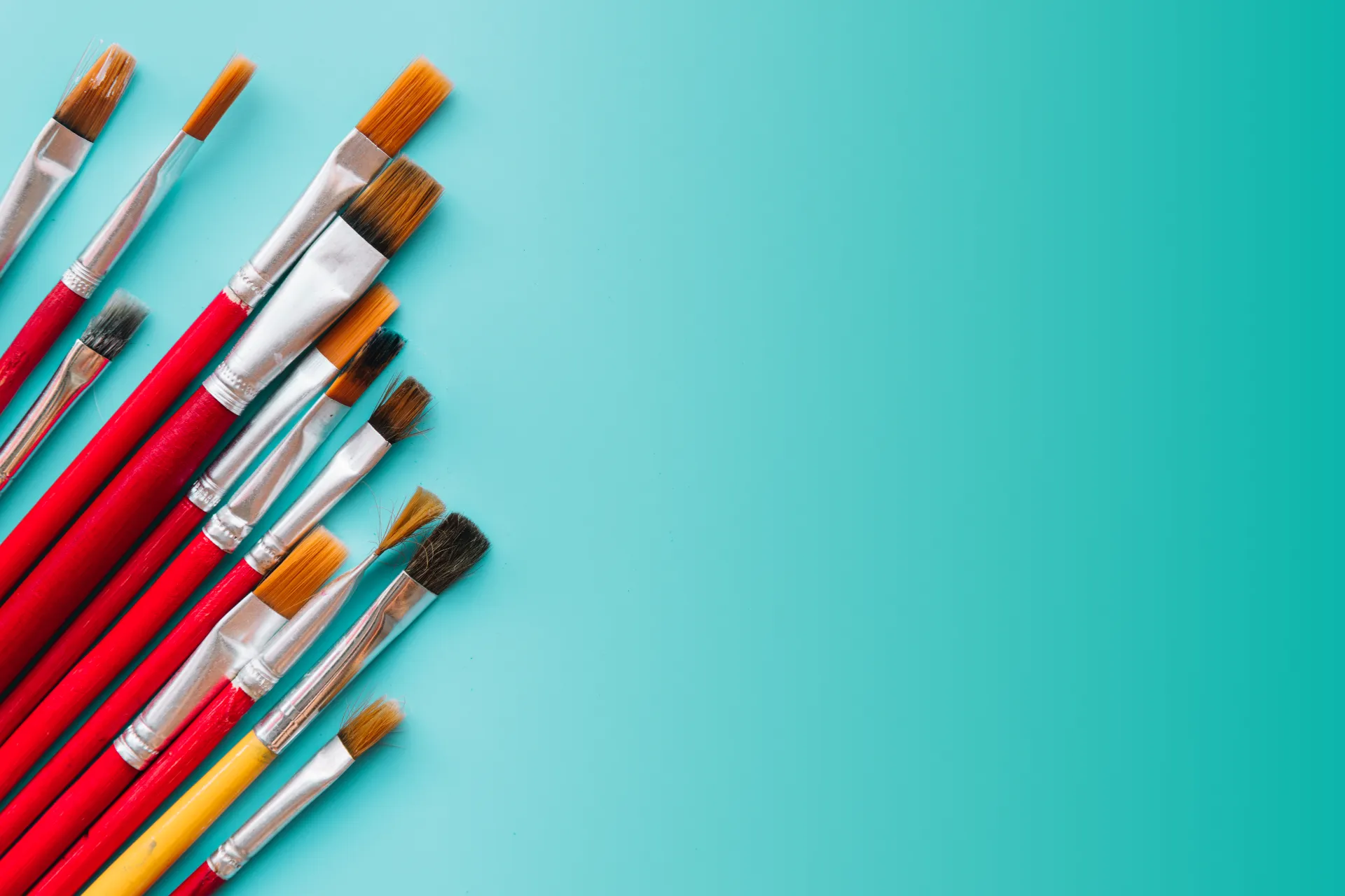 Zestaw różnych pędzli artystycznych z czerwonymi trzonkami ułożonych wzdłuż lewej krawędzi na turkusowym tle, zapraszający do wyboru i rezerwacji terminu w Studio Pan Kleks.