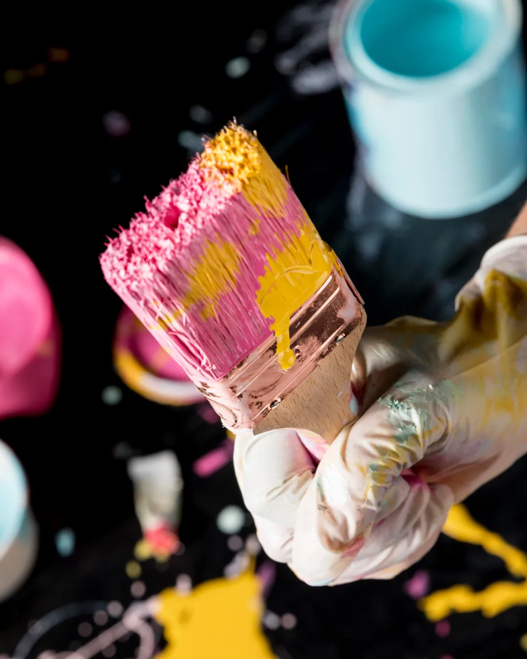 Dłoń w białej rękawiczce ubrudzonej farbą trzyma szeroki pędzel z różową i żółtą farbą na tle kolorowych puszek w pracowni Pan Kleks.