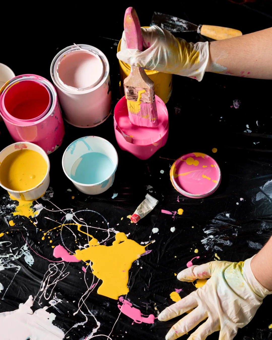Dłonie w rękawiczkach nad blatem pokrytym czarną folią z rozlaną kolorową farbą i otwartymi pojemnikami, ilustrujące wolność tworzenia bez konieczności sprzątania po sesji w Studio Pan Kleks.