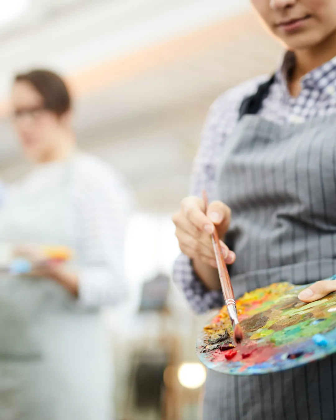 Uczestniczka warsztatów w szarym fartuchu trzyma paletę z wymieszanymi farbami i pędzel, przygotowując się do malowania w studio.